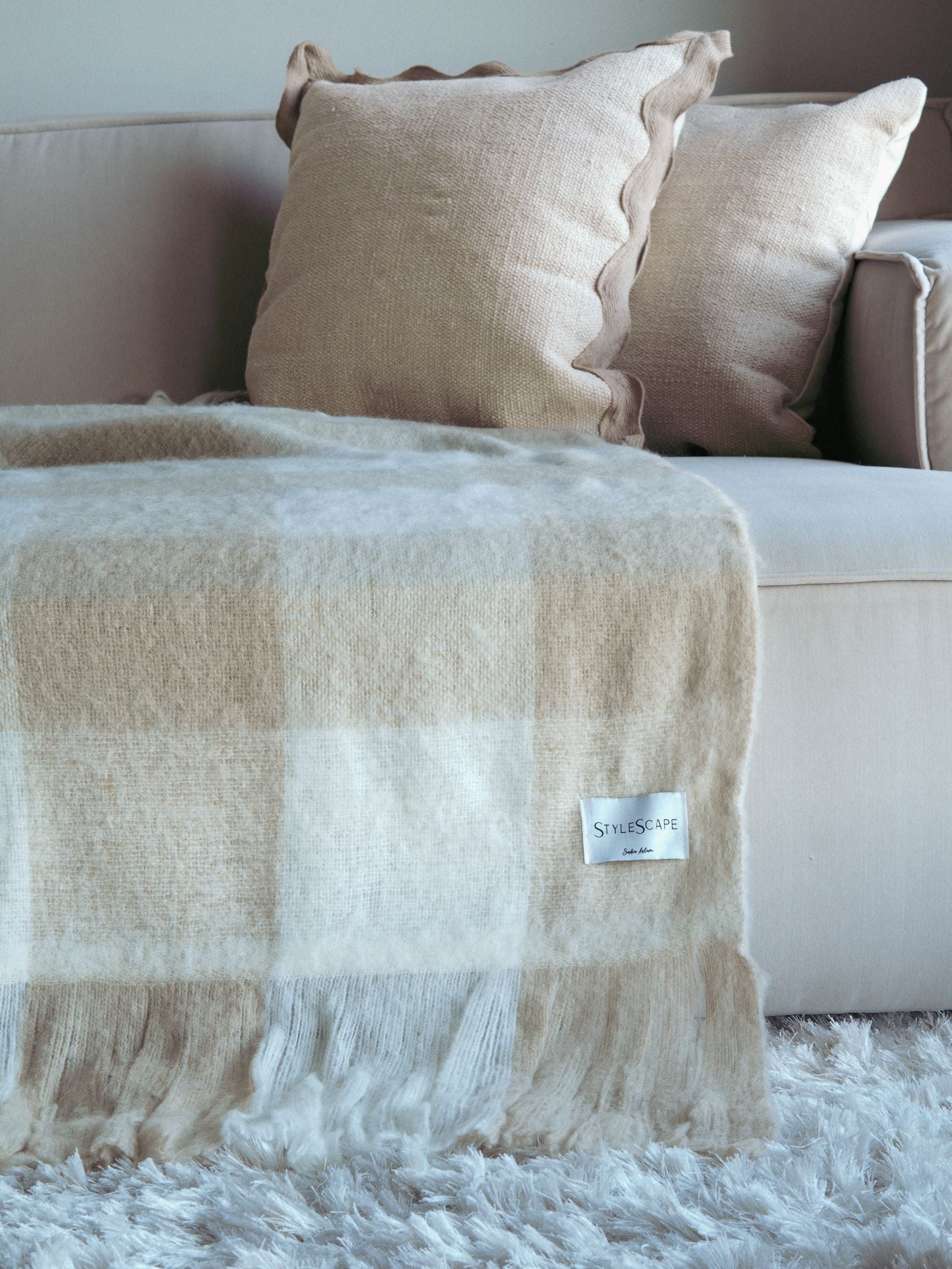 Et teppe i beige og off-white fargevalg, plassert på en seng med to puter oppå, i et rom med lyst interiør.